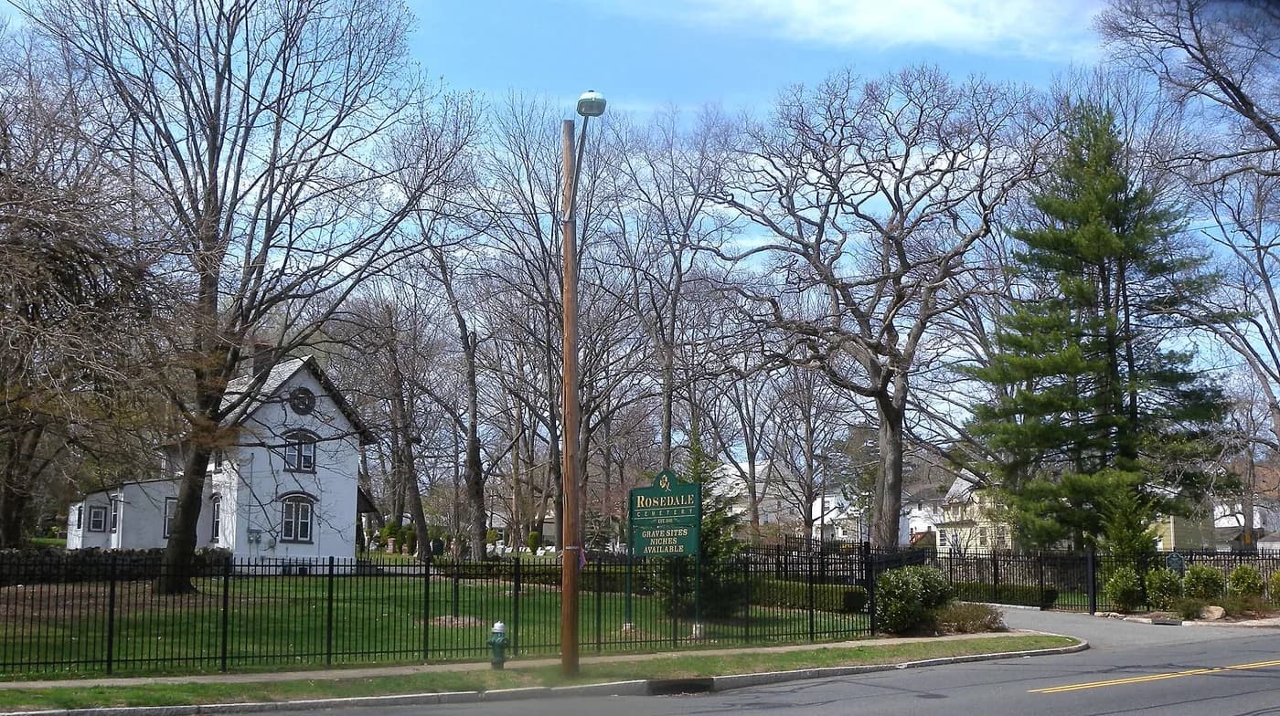 The edge of Rosedale Cemetery, with its fence, sign and tall trees.