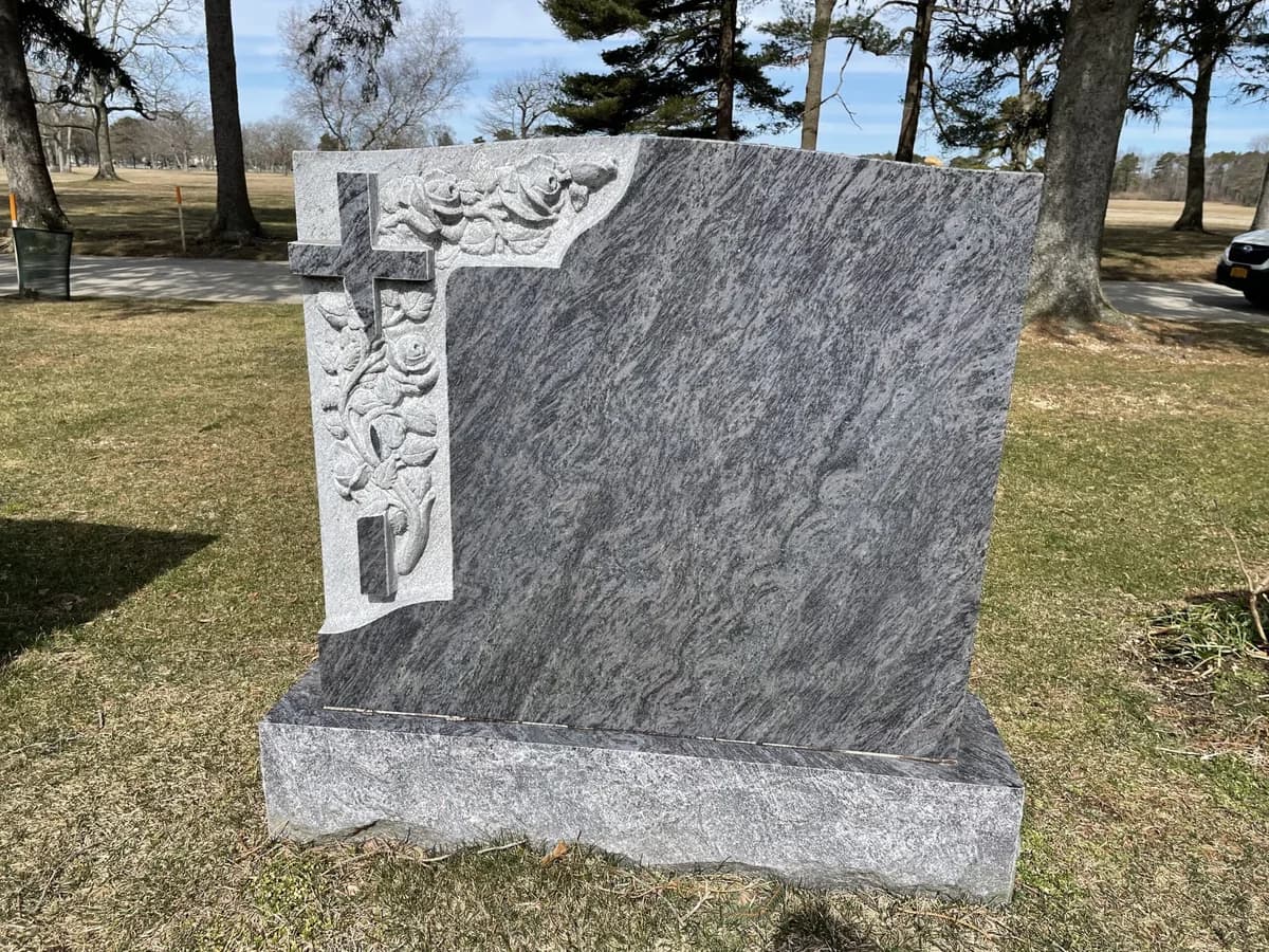 Grey granite headstone with a sculpted cross and foliage standing proud of a stepped corner.